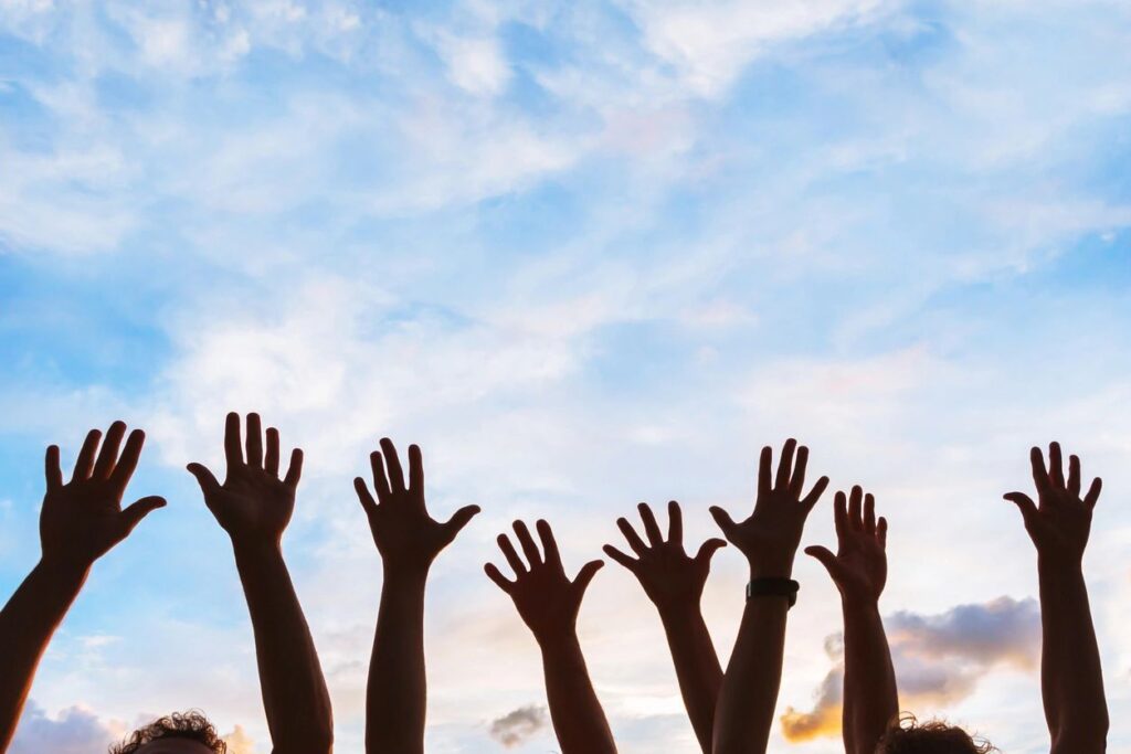 Raised hands reaching towards a bright blue sky with clouds.
